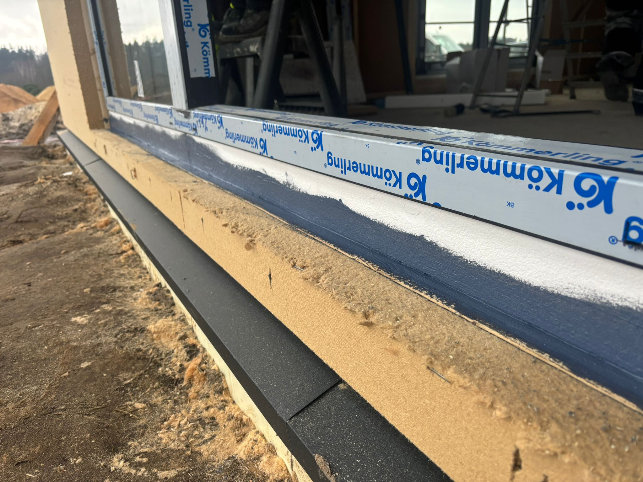 Widok z bliska na montaż okna, widoczna warstwa izolacyjna z pianki, listwa okienna z logo Kömmmerling i świeżo pomalowana rama okienna w kolorze ciemnoniebieskim.