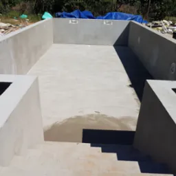 Surowy basen z betonu ze schodami, w trakcie budowy, z widocznymi elementami konstrukcyjnymi i wykończeniowymi. Widok z góry na wnętrze niecki basenowej.