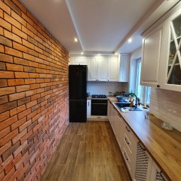 Majsterhome - Wnętrze kuchni po remoncie: ściana z cegły, drewnopodobne płytki na podłodze, białe meble kuchenne i czarna lodówka. Widok na całe, wąskie pomieszczenie.