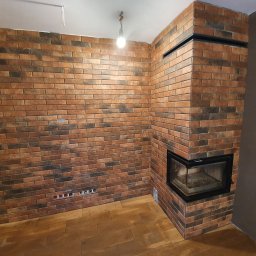Majsterhome - Wykończony kominek z cegły klinkierowej i ściana w tym samym stylu w Leoncin. Widoczne gniazdka elektryczne i drewniana podłoga.