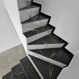 Wykończone schody z czarno-białych płyt marmurowych, widok z góry, z fragmentem surowej betonowej podłogi. Minimalistyczne połączenie bieli ścian i wzoru kamienia.