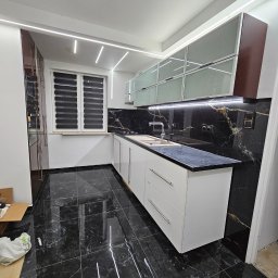 Majsterhome - Nowoczesna kuchnia z czarnymi marmurowymi płytkami podłogowymi i ściennymi, białymi szafkami i oświetleniem LED. Widok na okno z żaluzjami.