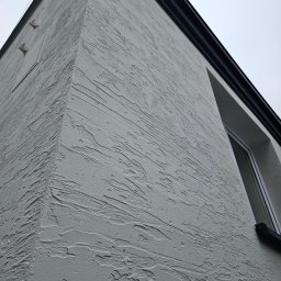 Majsterhome - Ściana budynku z nową, jasnoszarą strukturą tynku barankowego, widoczna z dołu pod kątem, z fragmentem okna i czarnym wykończeniem dachu.