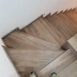 Majsterhome - Ułożone drewnopodobne panele na schodach, widok z góry na stopy w butach sportowych. Realizacja wykończenia schodów z dbałością o detale.