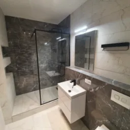 Nowoczesna łazienka z kabiną typu walk-in, umywalką na szafce i marmurowymi płytkami w odcieniach szarości. Minimalistyczny design, eleganckie wykończenie.