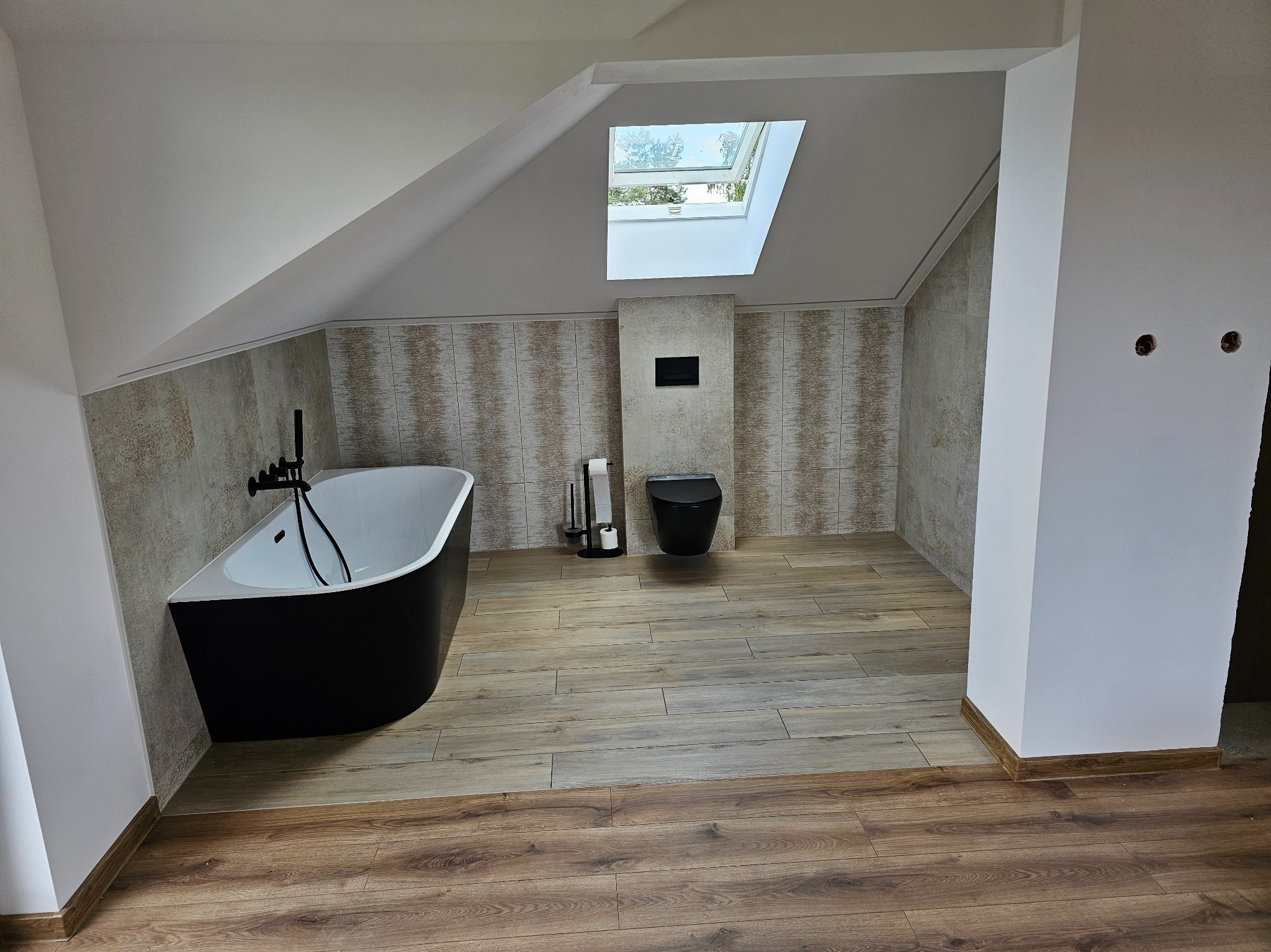 Wykończona łazienka na poddaszu z czarną wanną i toaletą. Beżowe płytki imitujące kamień na ścianach, drewnopodobne panele na podłodze, okno dachowe.
