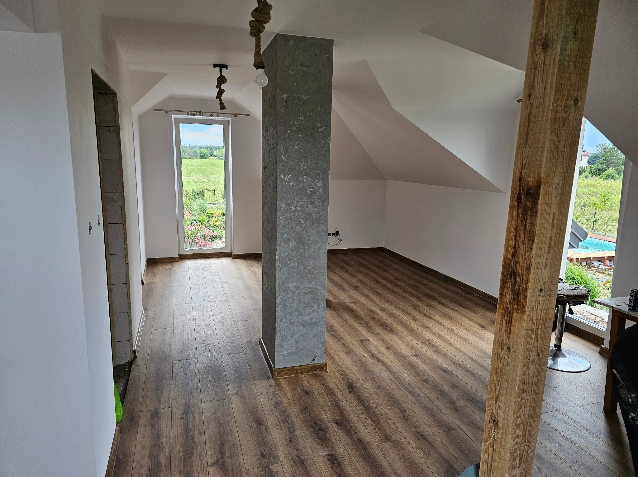 Jasne poddasze z drewnianą podłogą, kolumna wykończona w szarości, okno z widokiem na zieleń. Częściowo wykończone wnętrze w stylu rustykalnym.