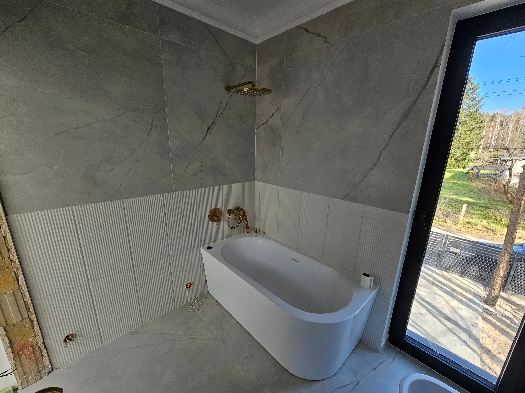 Nowoczesna łazienka z wanną, szarymi płytkami imitującymi marmur i złotą armaturą. Duże okno z widokiem na zewnątrz. Częściowo wykończona ściana z płytkami.
