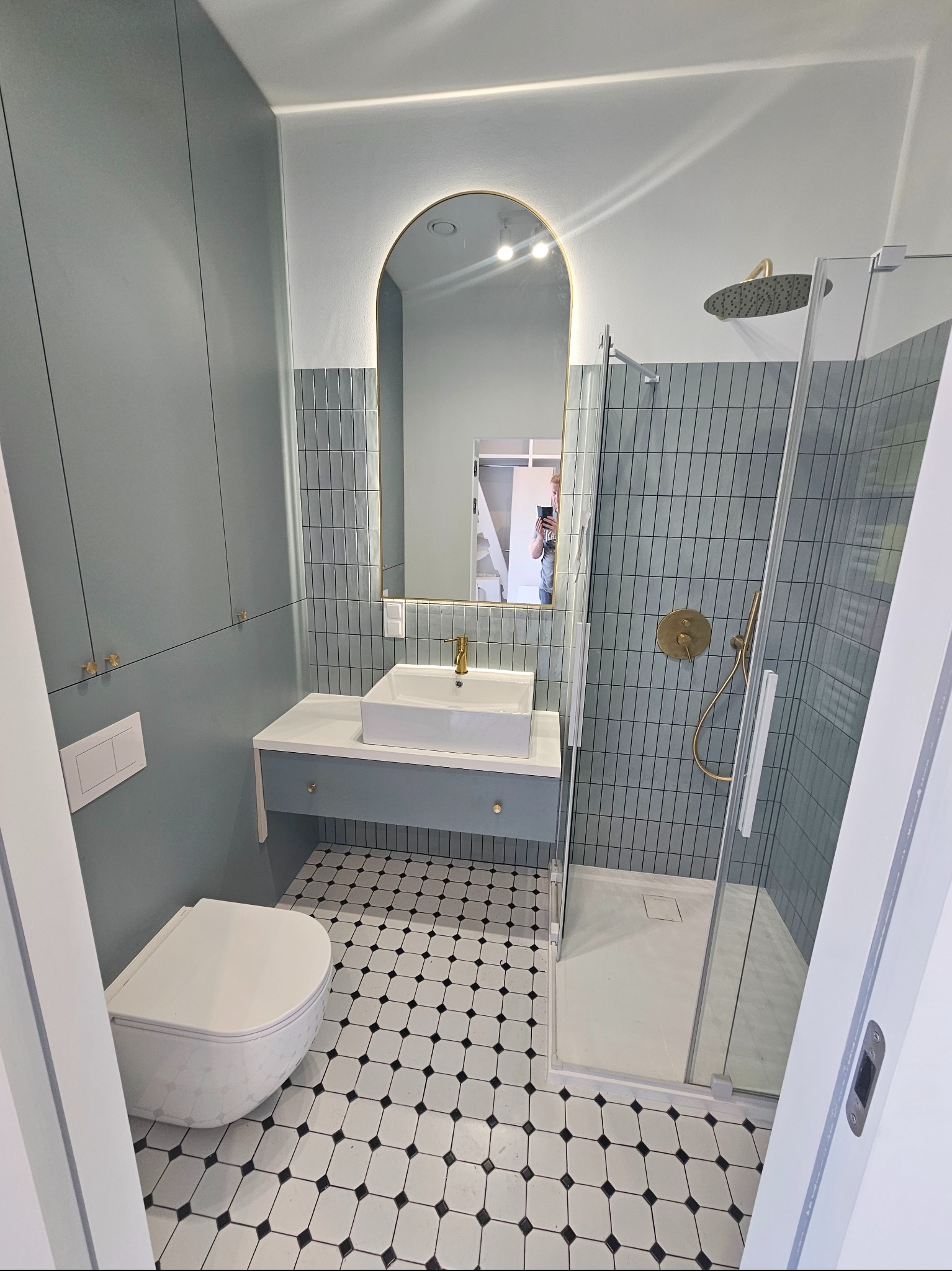 Wykończona łazienka w stylu retro z kabiną prysznicową, umywalką na szafce, toaletą i czarno-białą podłogą. Na ścianach szaro-błękitne płytki i szafki.