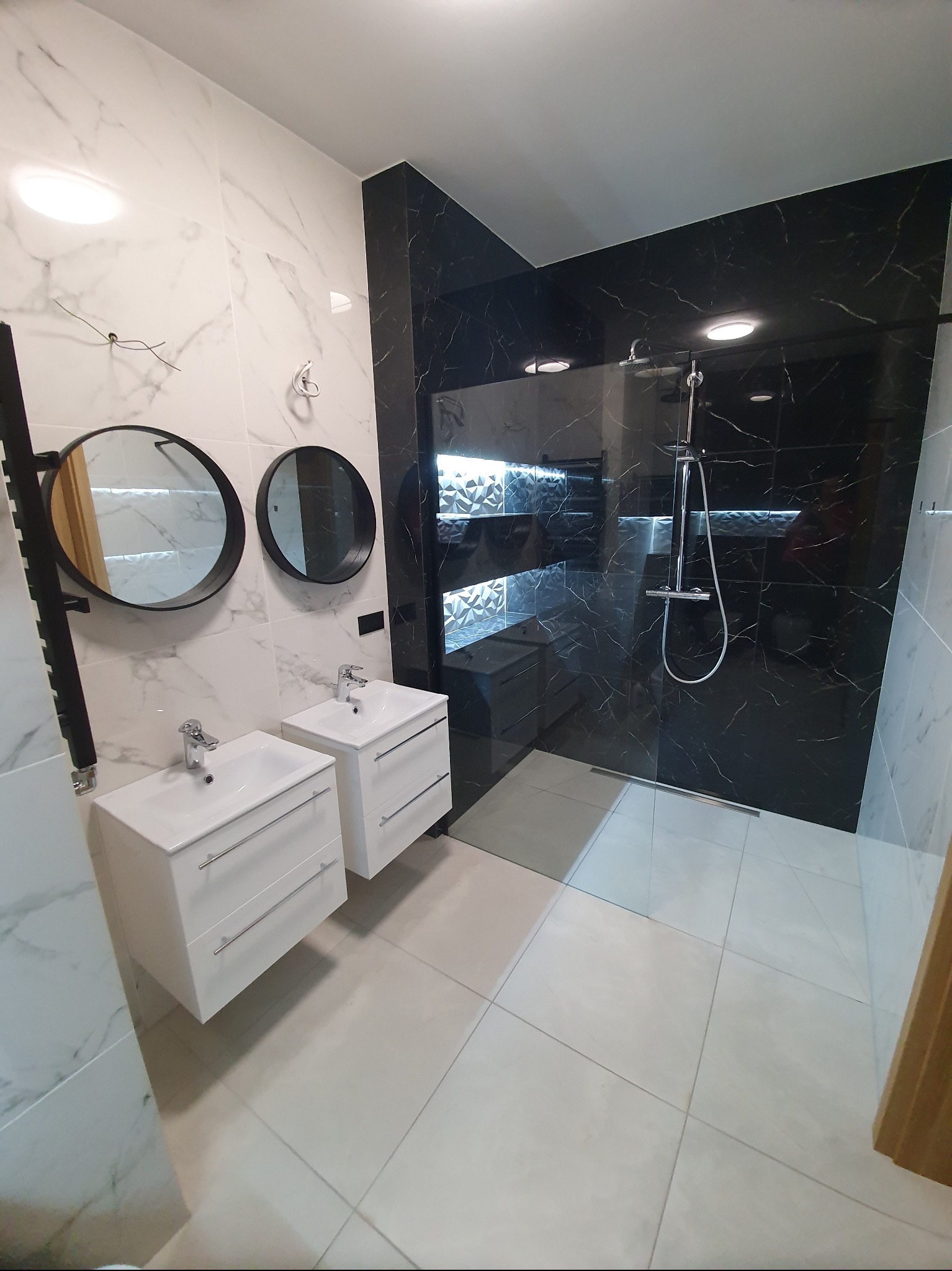 Nowoczesna łazienka z czarno-białymi marmurkowymi płytkami, dwoma umywalkami z szafkami i okrągłymi lustrami, kabina prysznicowa walk-in.