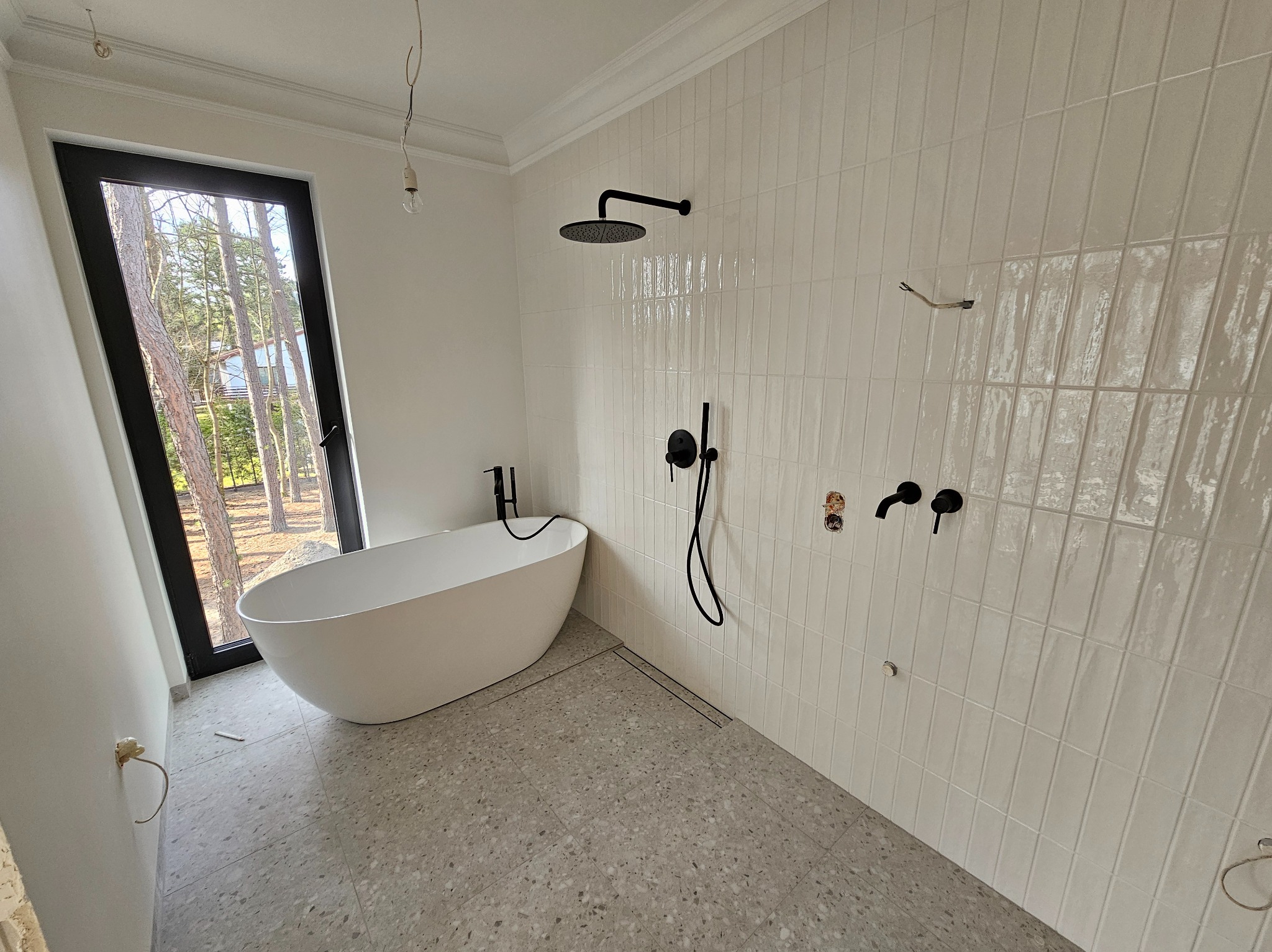 Nowoczesna łazienka w trakcie wykańczania: białe płytki na ścianach, wolnostojąca wanna, czarna armatura i okno z widokiem na drzewa. Minimalistyczny design.