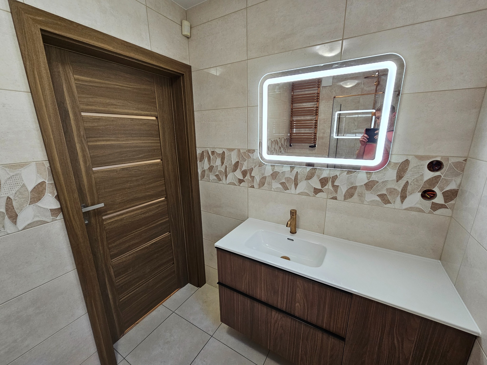 Nowoczesna łazienka po remoncie:  brązowe drzwi z poziomymi frezami, szafka pod umywalkę z drewnopodobnym frontem, podświetlane lustro.