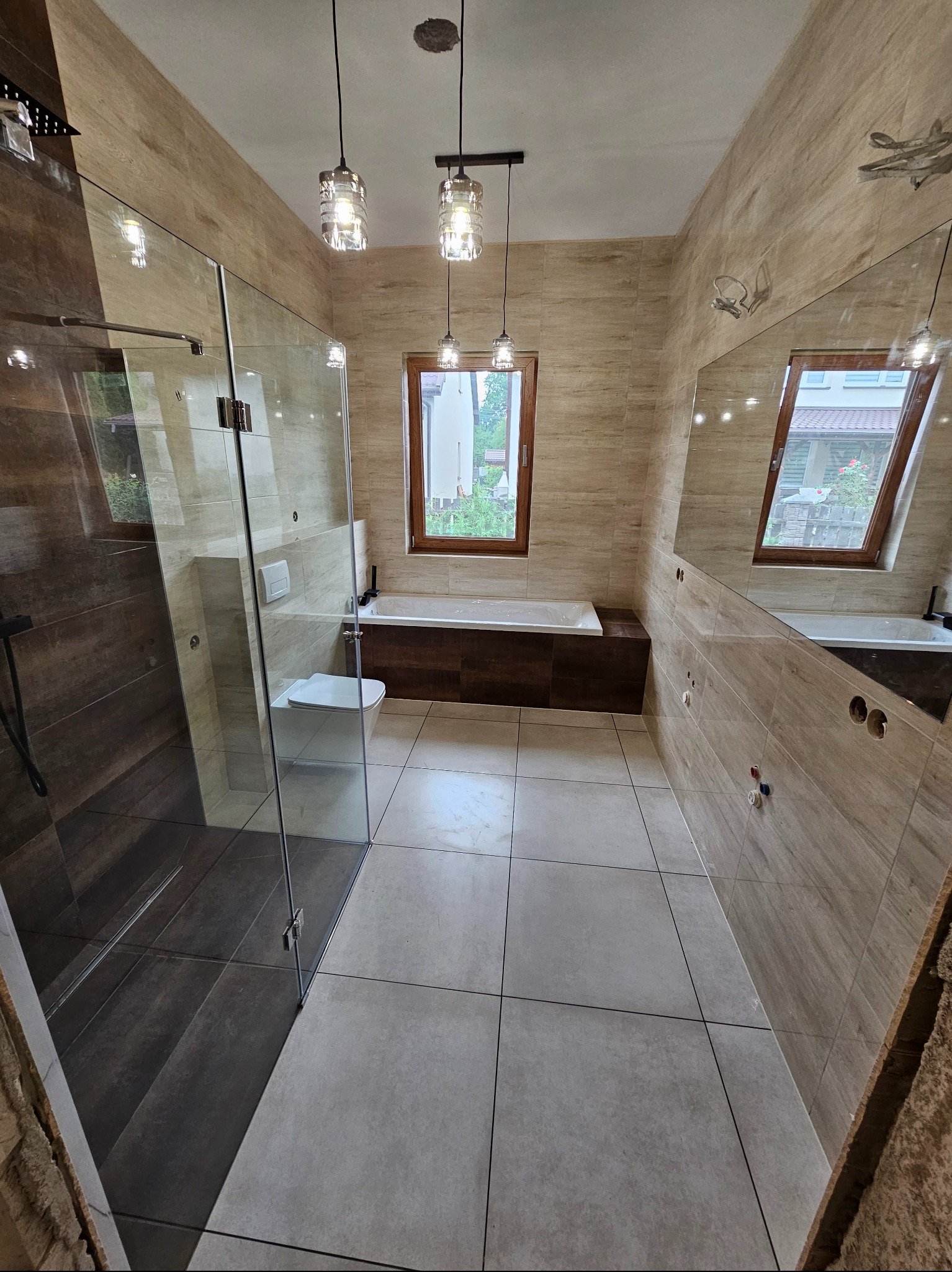 Nowoczesna łazienka z kabiną prysznicową typu walk-in, beżowymi płytkami imitującymi drewno i wiszącymi lampami z przezroczystego szkła.