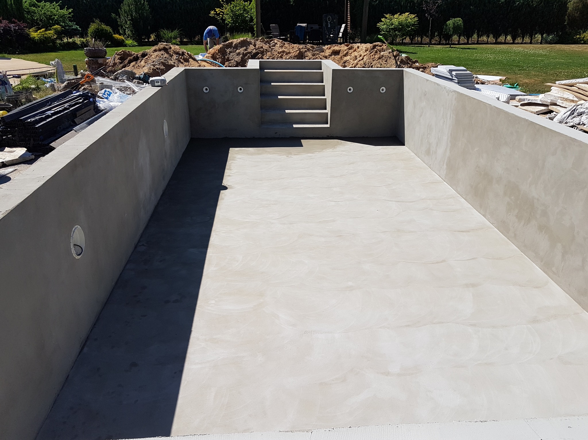 Surowa betonowa konstrukcja basenu w ziemi z wbudowanymi schodami, przygotowana do dalszych prac wykończeniowych, z widocznymi instalacjami i otoczeniem budowy.