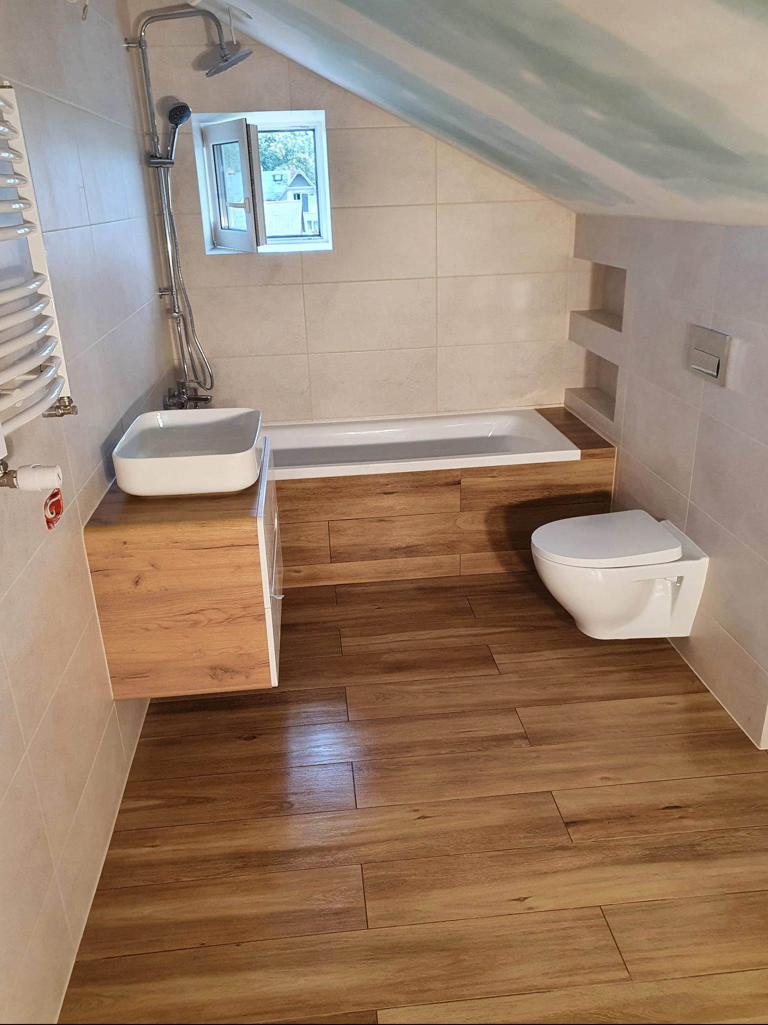 Wykończona łazienka na poddaszu z drewnopodobną podłogą, białą armaturą i beżowymi płytkami. Umywalka nablatowa na szafce z drewnianym frontem.