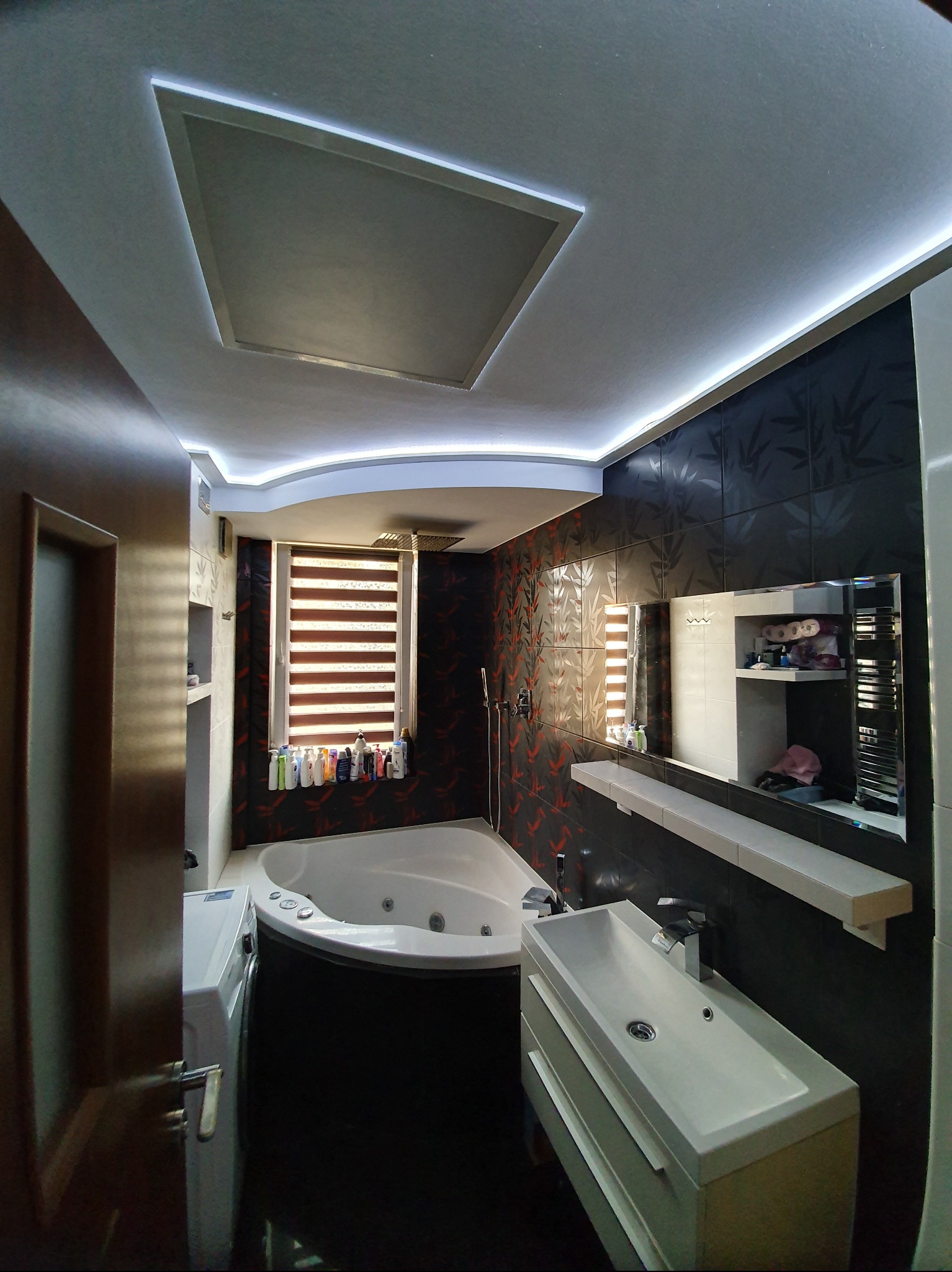 Nowoczesna łazienka z wanną z hydromasażem, czarnymi płytkami z motywem roślinnym i podświetleniem LED sufitu. Umywalka z szafką i lustro dopełniają aranżację.