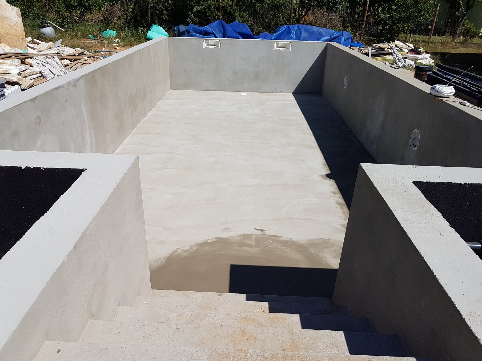 Surowy basen z betonu ze schodami, w trakcie budowy, z widocznymi elementami konstrukcyjnymi i wykończeniowymi. Widok z góry na wnętrze niecki basenowej.
