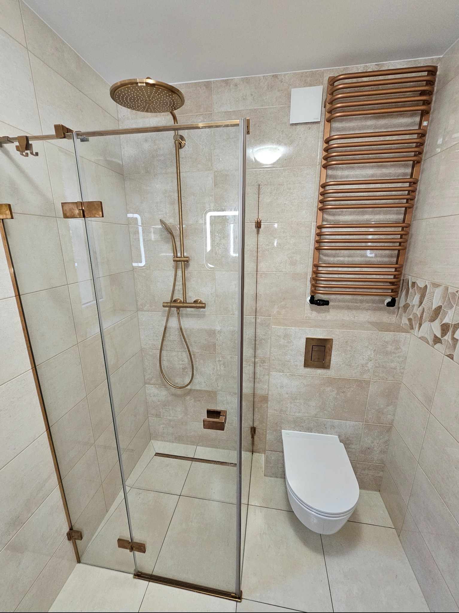 Beżowa łazienka z kabiną prysznicową typu walk-in z złotymi okuciami, grzejnikiem drabinkowym w kolorze miedzianym i podwieszaną toaletą. Estetyczne wykończenie.