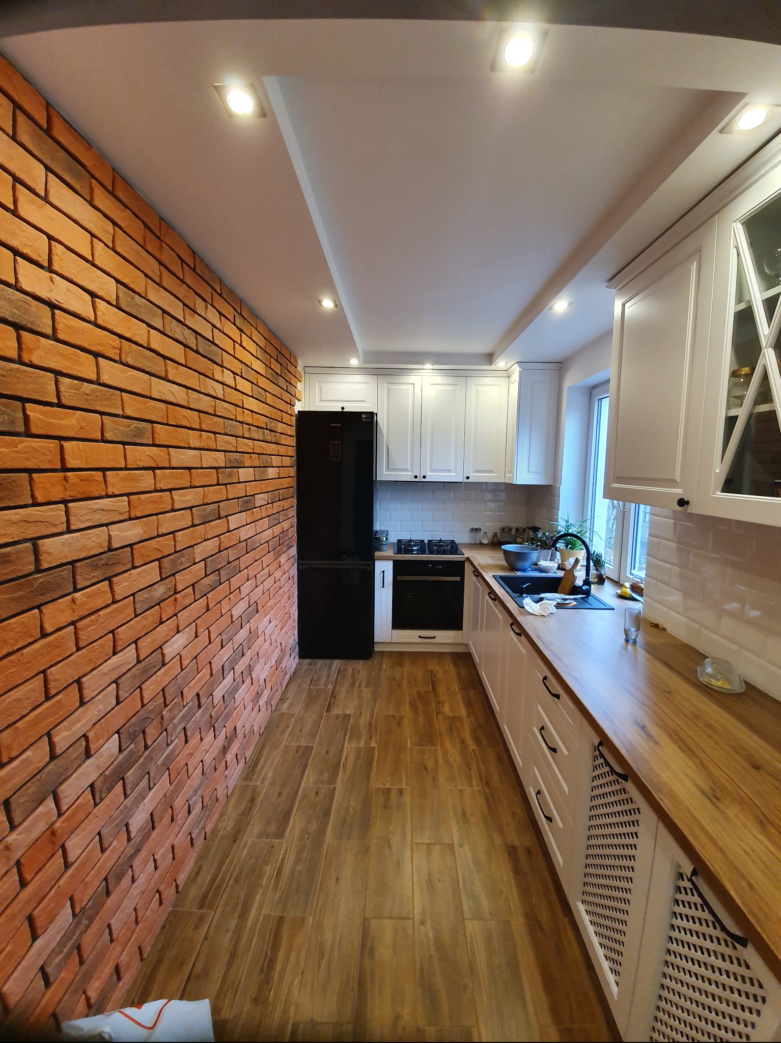 Wnętrze kuchni po remoncie: ściana z cegły, drewnopodobne płytki na podłodze, białe meble kuchenne i czarna lodówka. Widok na całe, wąskie pomieszczenie.