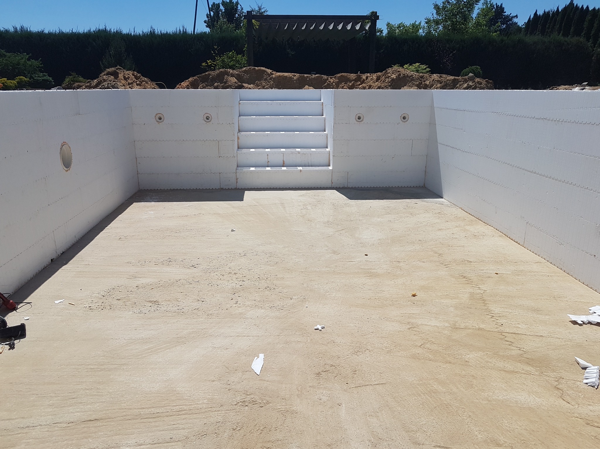 Niedokończony basen z białymi ścianami i schodami, widok z góry, otoczony zielenią i konstrukcją z drewna. Dno basenu pokryte piaskiem.