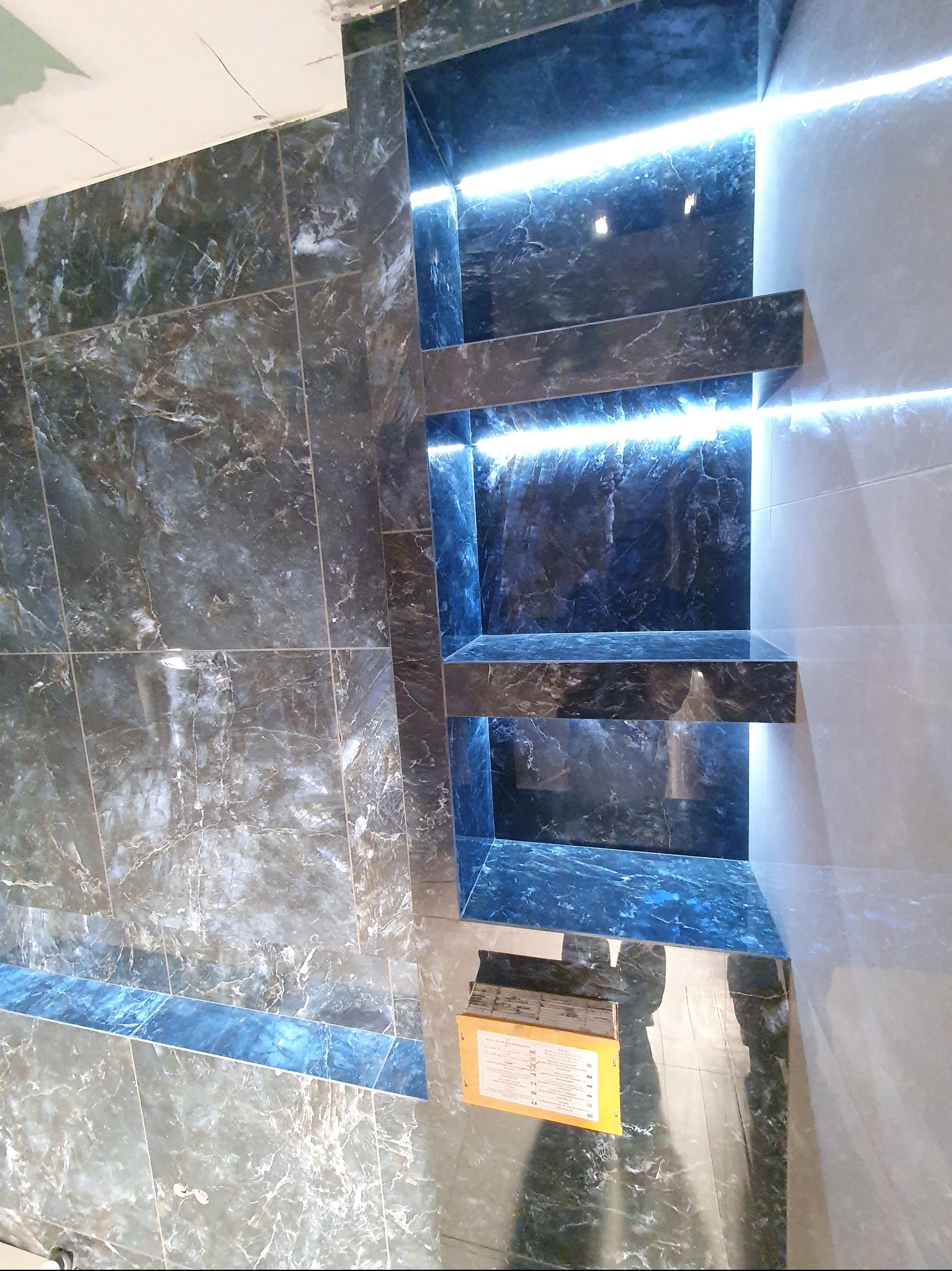 Wykończona łazienka z ciemnymi marmurkowymi płytkami i podświetlanymi półkami wnękowymi. Nowoczesny design, widok na fragment podłogi i ścian.