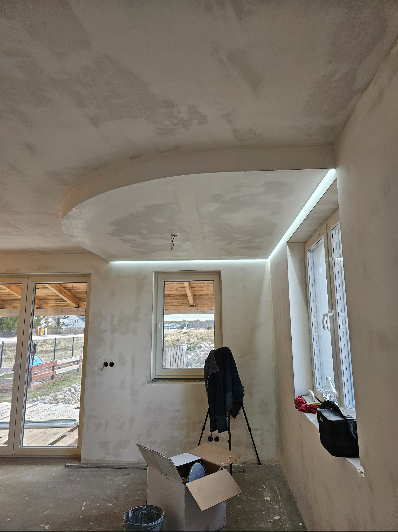 Wnętrze w trakcie szpachlowania: sufit z owalną wnęką i oświetleniem LED, ściany w szpachli, okna, drzwi balkonowe, narzędzia pracy, karton na podłodze.