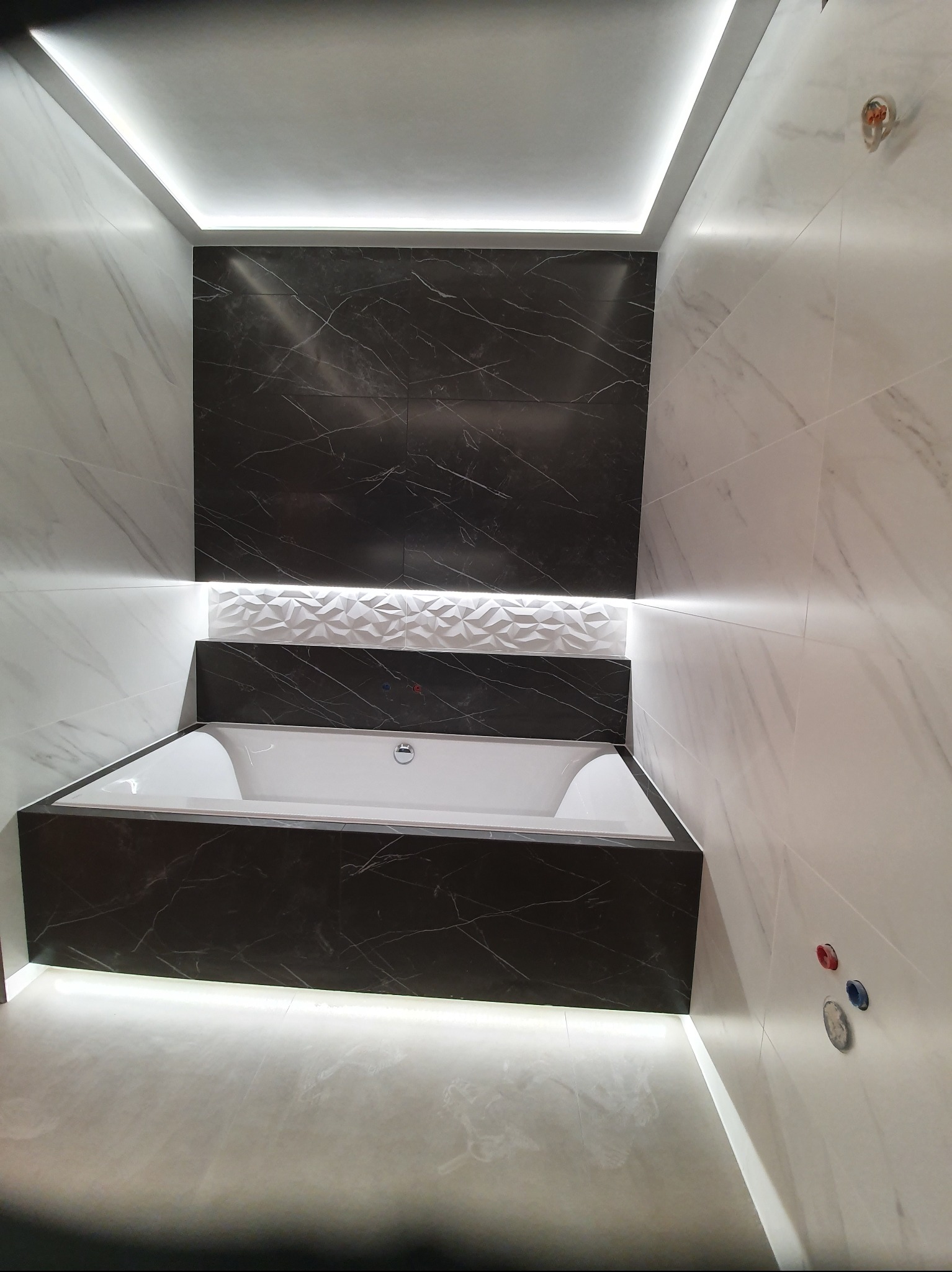 Nowoczesna łazienka z wanną obudowaną czarnym marmurem, białe płytki imitujące marmur, oświetlenie LED podkreślające geometrię wnętrza.