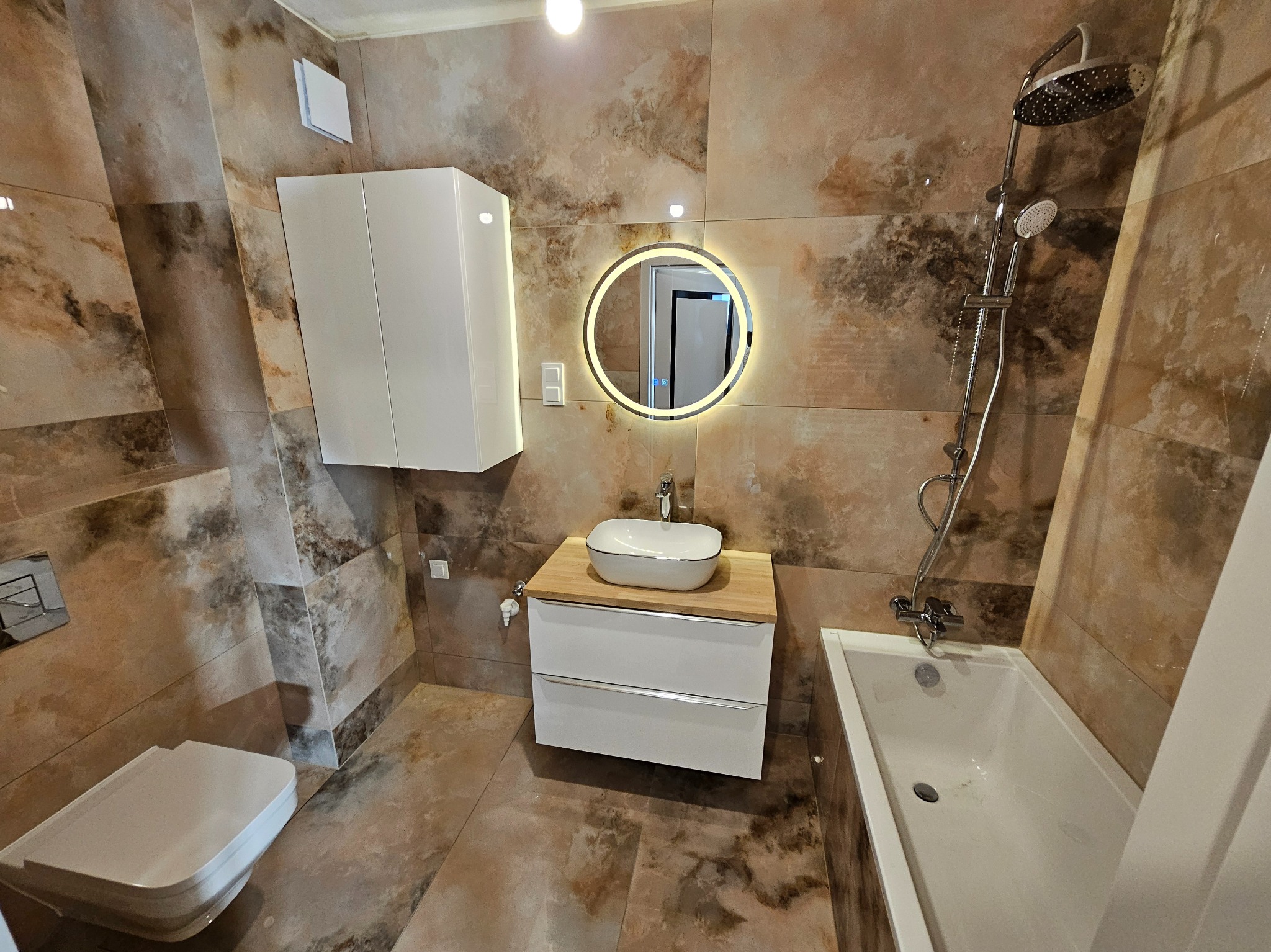 Nowoczesna łazienka z beżowymi płytkami imitującymi marmur, białą armaturą i okrągłym lustrem z podświetleniem LED. Widoczna umywalka nablatowa i wanna.