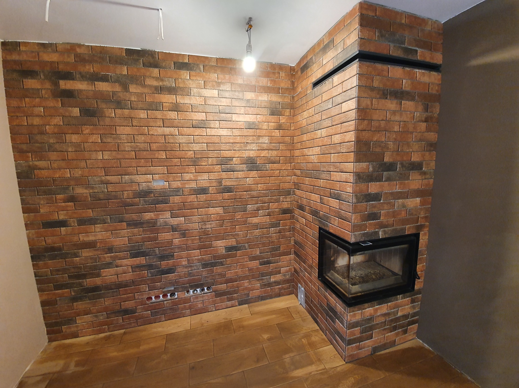 Wykończony kominek z cegły klinkierowej i ściana w tym samym stylu w Leoncin. Widoczne gniazdka elektryczne i drewniana podłoga.