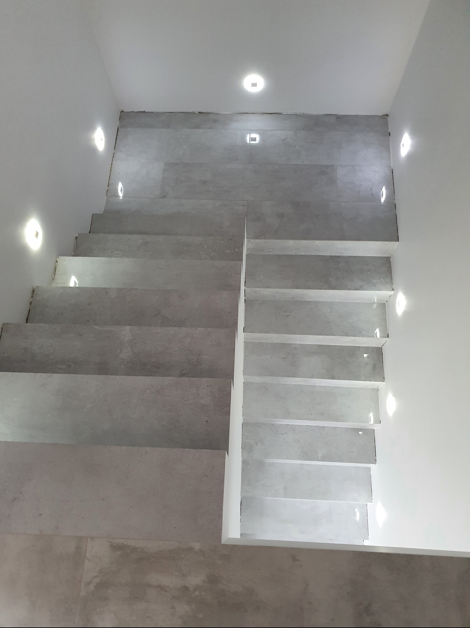 Wykończone schody z szarych płytek, oświetlone punktowymi lampami w ścianach i suficie. Minimalistyczny design, widok z góry.