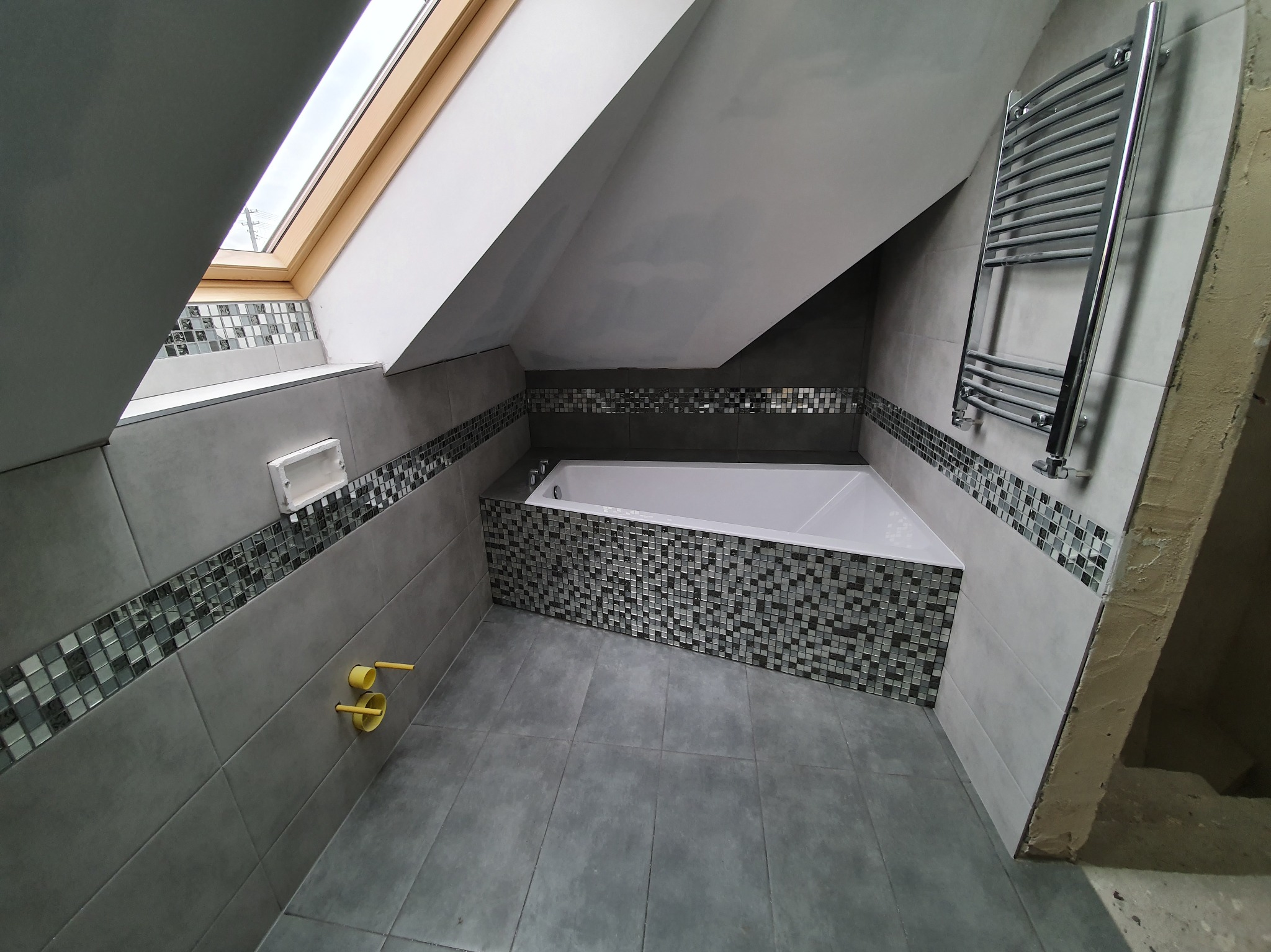 Szara łazienka na poddaszu z mozaiką, wanną i grzejnikiem drabinkowym. Okno dachowe wpuszcza światło. Widać fragment ściany w stanie surowym.