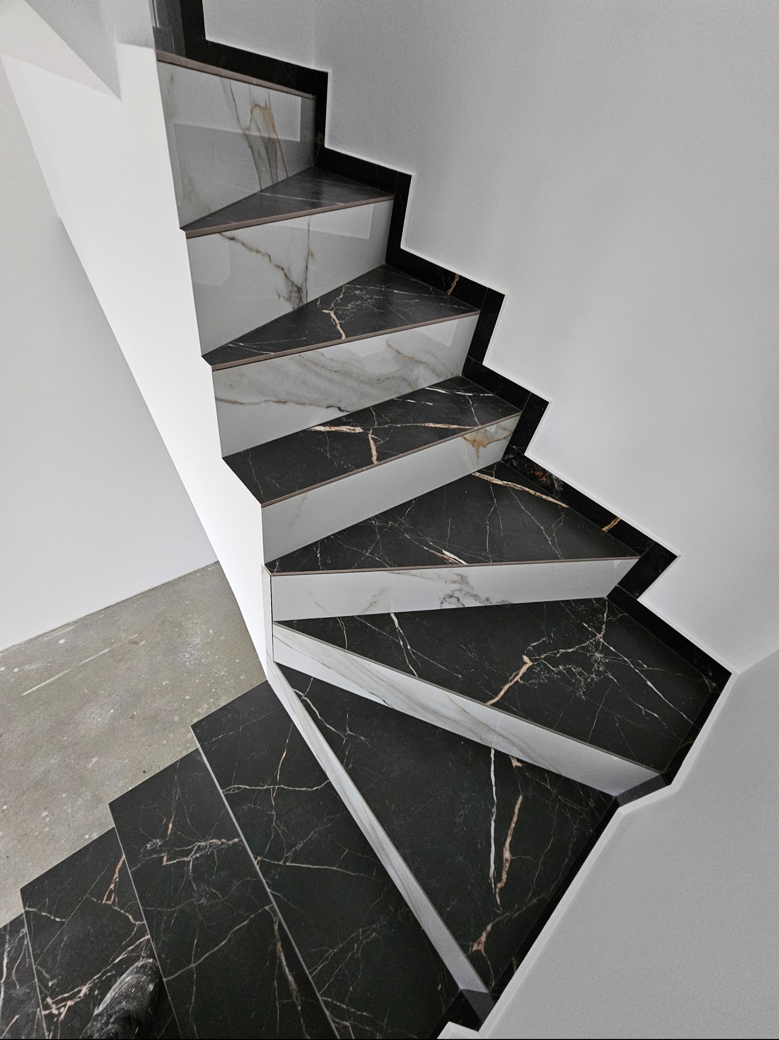 Wykończone schody z czarno-białych płyt marmurowych, widok z góry, z fragmentem surowej betonowej podłogi. Minimalistyczne połączenie bieli ścian i wzoru kamienia.