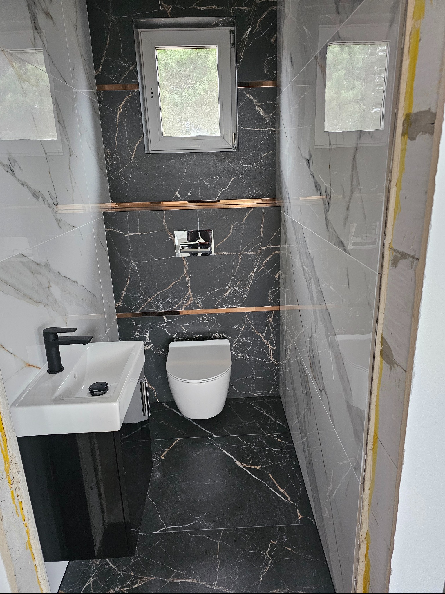 Nowoczesna łazienka z czarno-białymi płytkami imitującymi marmur, złotymi akcentami i wiszącą toaletą. Widoczne wykończenia i okno.