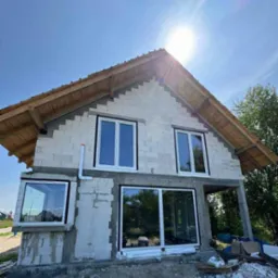 Surowy stan domu z betonu komórkowego w trakcie budowy. Widoczne białe okna, drewniana konstrukcja dachu i instalacja odprowadzająca wodę. Słoneczny dzień.