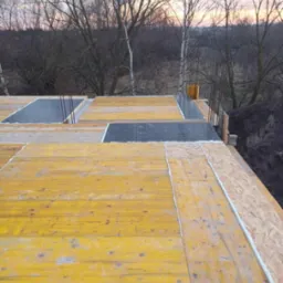 Szlifowana sklejka szalunkowa, zbrojenie i pianka montażowa na budowie. Widok z góry na konstrukcję w trakcie wznoszenia, z drzewami w tle o zmierzchu.