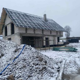 CORNELL CONSTRUCTION - KAROLINA BIELICKA - Stan surowy otwarty domu z białego pustaka, z częściowo położonym dachem i kominem. Na pierwszym planie hałda ziemi i niebieski wąż. Zimowa sceneria, lekko ośnieżone.