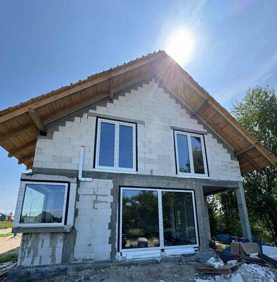 Surowy stan domu z betonu komórkowego w trakcie budowy. Widoczne białe okna, drewniana konstrukcja dachu i instalacja odprowadzająca wodę. Słoneczny dzień.