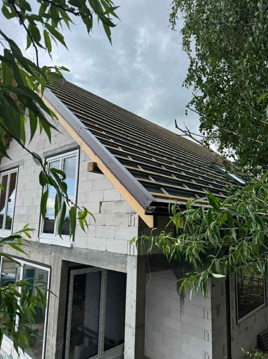 Budowa domu z białych bloczków: widoczne ściany, okna i konstrukcja dachu z łatami, ujęcie z dołu, częściowo zasłonięte przez liście drzew.