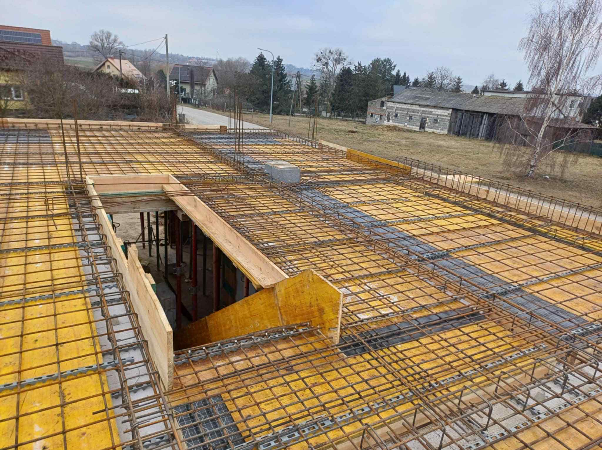 Zbrojenie stropu z żółtym szalunkiem na budowie domu jednorodzinnego. Widoczne stalowe pręty i elementy konstrukcyjne. W tle zabudowa wiejska i pochmurne niebo.