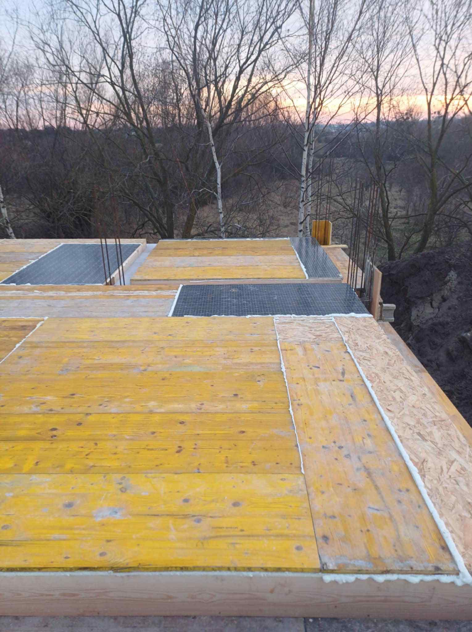 Szlifowana sklejka szalunkowa, zbrojenie i pianka montażowa na budowie. Widok z góry na konstrukcję w trakcie wznoszenia, z drzewami w tle o zmierzchu.