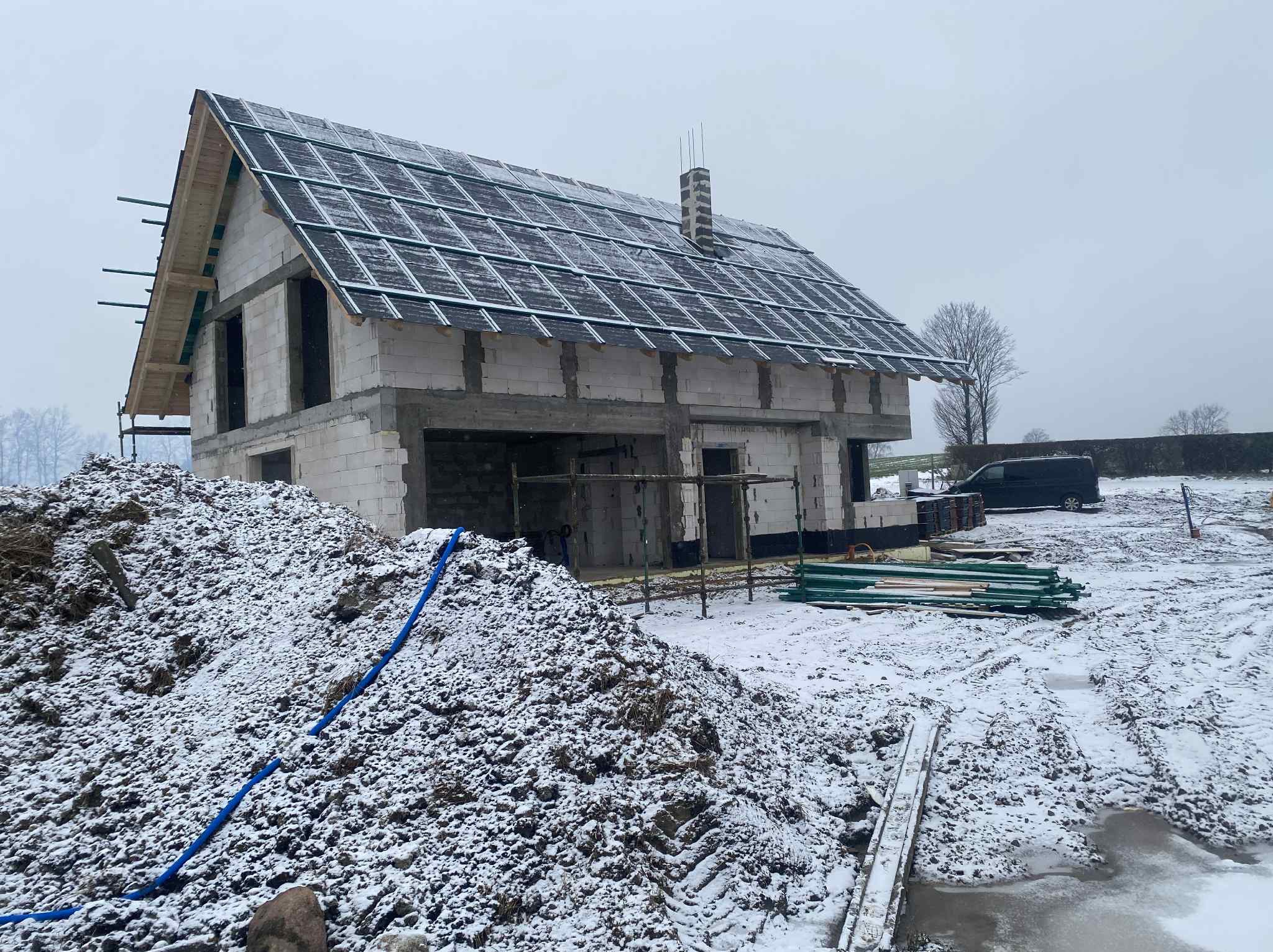 Stan surowy otwarty domu z białego pustaka, z częściowo położonym dachem i kominem. Na pierwszym planie hałda ziemi i niebieski wąż. Zimowa sceneria, lekko ośnieżone.