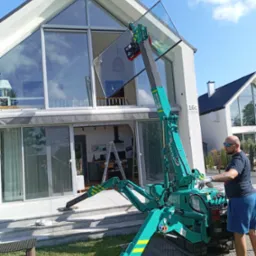 Montaż okna za pomocą mini żurawia Maeda na tle nowoczesnego domu z dużymi przeszkleniami.