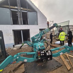 Nattex - Zielony mini dźwig pająk marki Maeda podnosi dużą szybę okienną na placu budowy domu z białych bloczków, w tle robotnicy w odblaskowych kamizelkach i niezadaszony budynek.