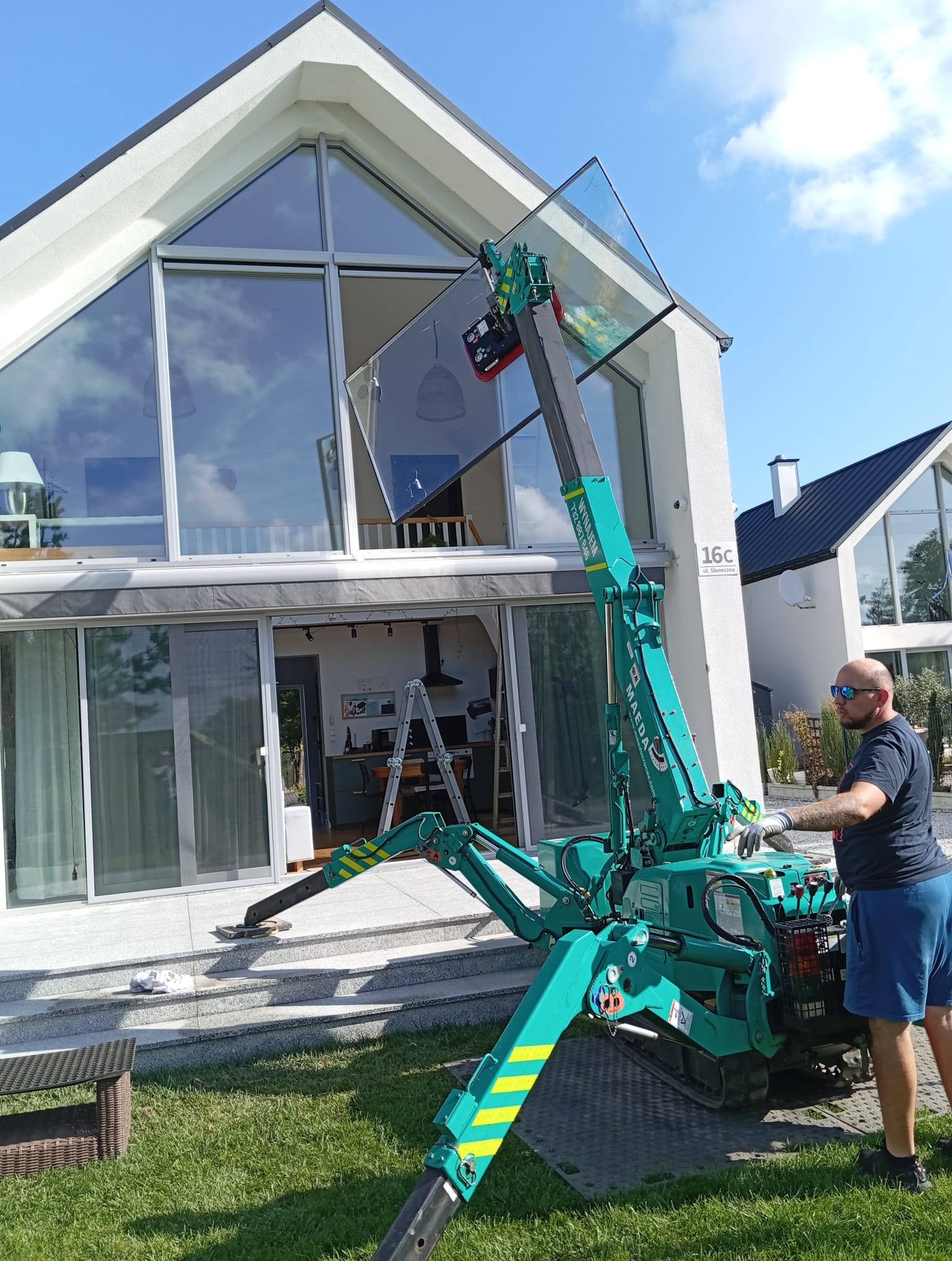 Montaż okna za pomocą mini żurawia Maeda na tle nowoczesnego domu z dużymi przeszkleniami.