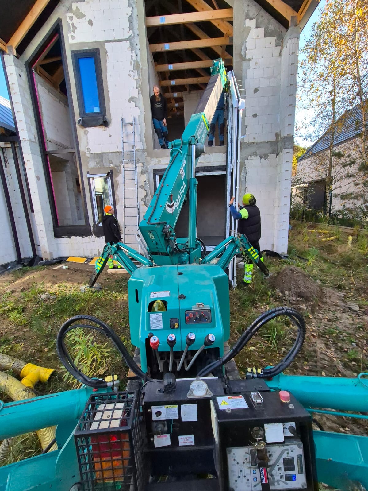 Kompaktowy żuraw Maeda w trakcie montażu okna w surowym stanie budynku z białego bloczka, operatorzy w kaskach i kamizelkach odblaskowych, widok z perspektywy operatora żurawia, częściowo widoczne...
