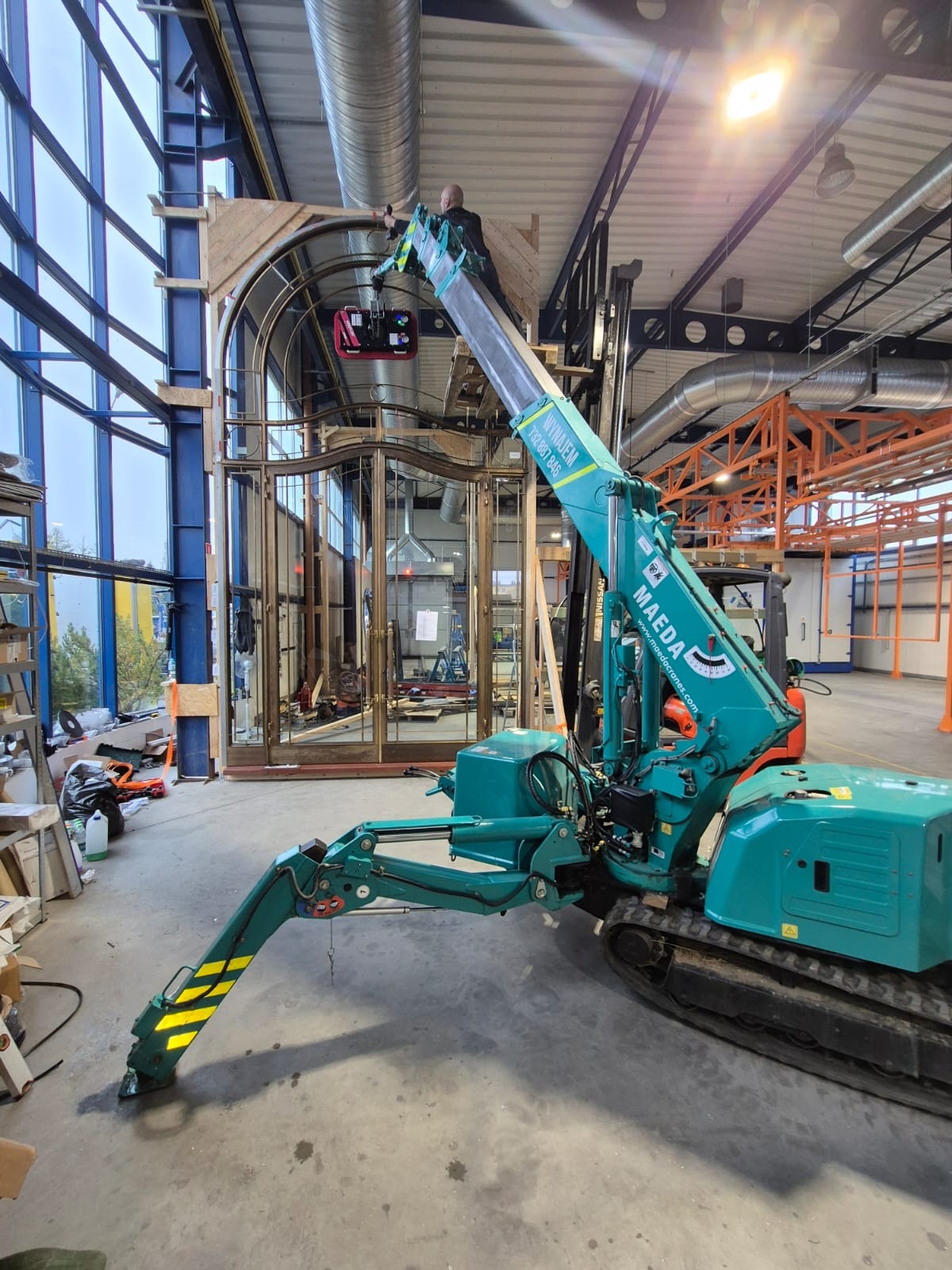 Kompaktowy, turkusowy dźwig gąsienicowy marki Maeda w hali, podnosi element konstrukcyjny z drewna i metalu, pracownik na platformie dźwigu, duże okna w tle.