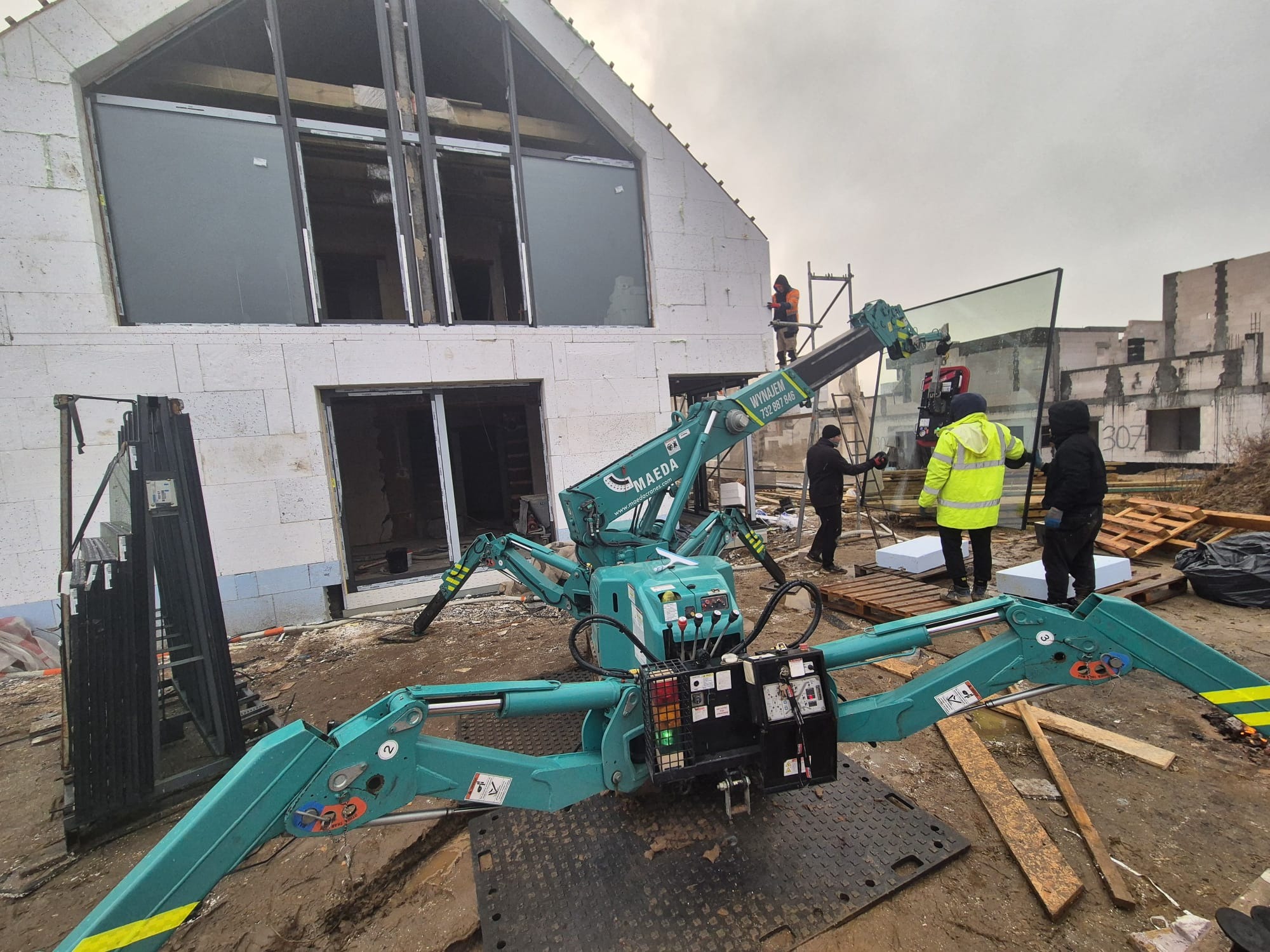 Zielony mini dźwig pająk marki Maeda podnosi dużą szybę okienną na placu budowy domu z białych bloczków, w tle robotnicy w odblaskowych kamizelkach i niezadaszony budynek.