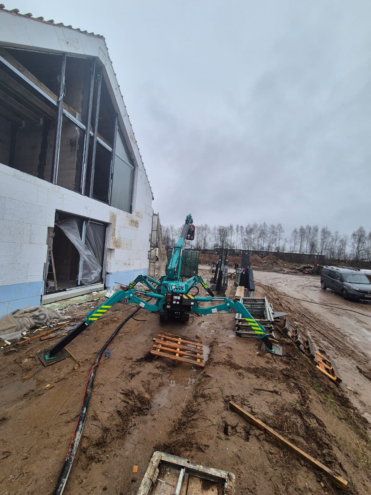 Kompaktowy dźwig pająk w kolorze turkusowym, rozstawiony na błotnistym terenie budowy, z widoczną surową konstrukcją budynku w tle i zaparkowanym samochodem dostawczym.