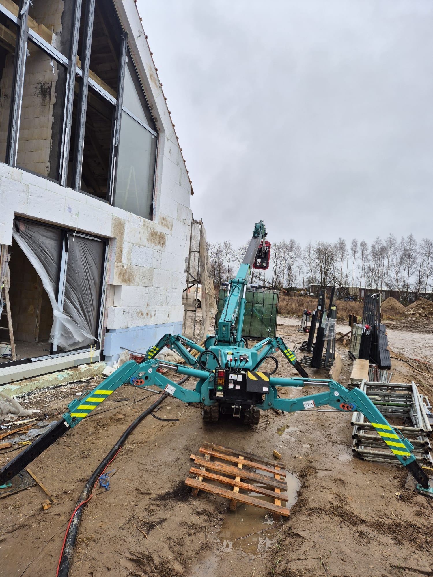 Niebiesko-zielony pająk-dźwig z żółtymi pasami, ustawiony na błotnistym placu budowy, podnosi ciężkie okno do budynku w stanie surowym.