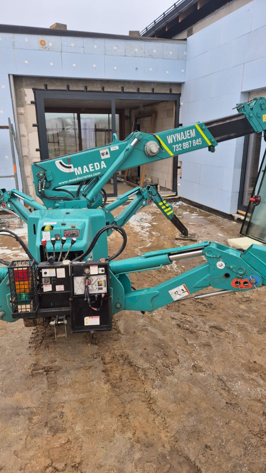 Mały, zielony żuraw Maeda z wysięgnikiem ustawionym przy oknie budynku w trakcie montażu, widoczne ślady gąsienic na błotnistym podłożu.